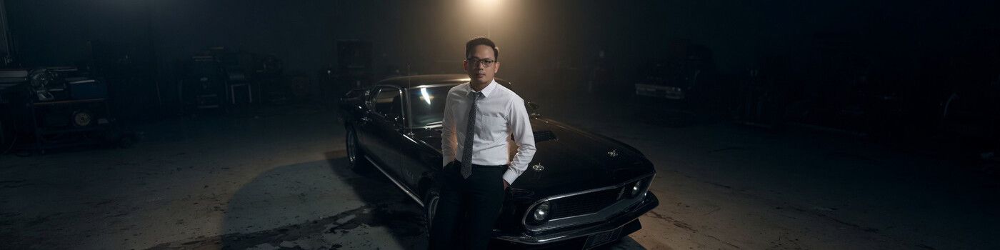 Anderson So in a white shirt and tie, leaning on a black classic Mustang in a dark garage